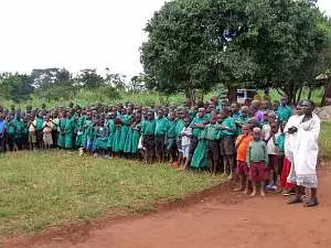 A large group of children, girls mainly in green dresses and boys in green shirts and shorts, line up for a welcome reception at Mugungulu School