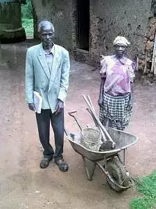 A man and wom stand next to a wheel barrow containing assorted farming implements